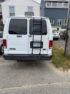 Rear view of a white Ford van, configured like an RV rental with a ladder and roof vent, parked on a street.