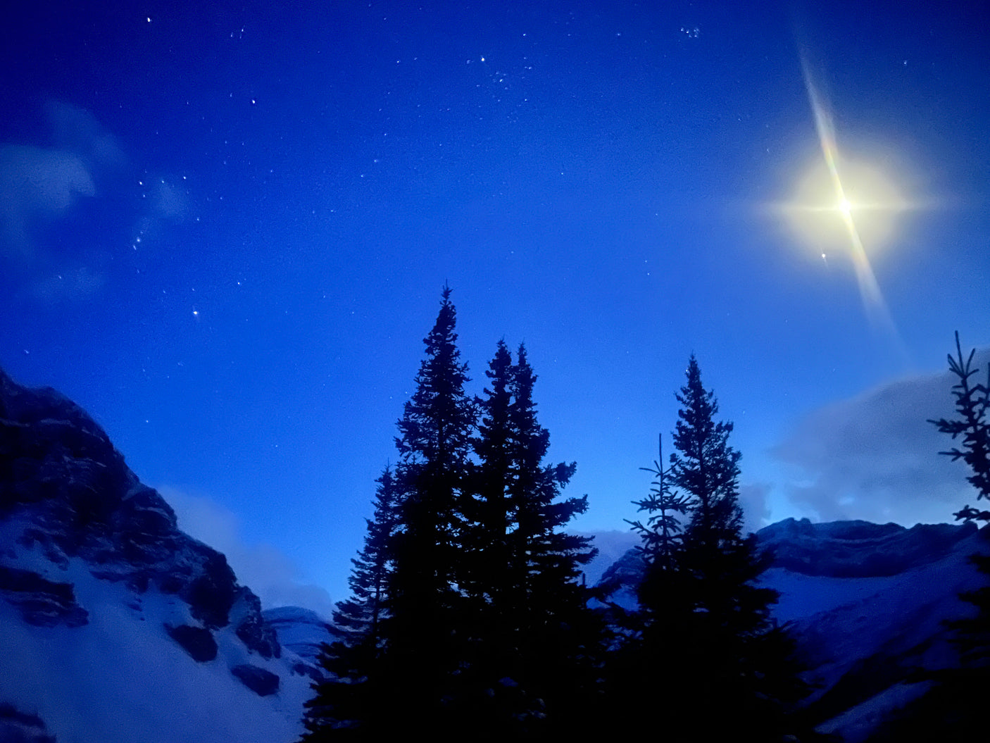 Deep blue night sky, sparkling stars, & glowing celestial object above snowy mountains & silhouetted pines. RV rental views!