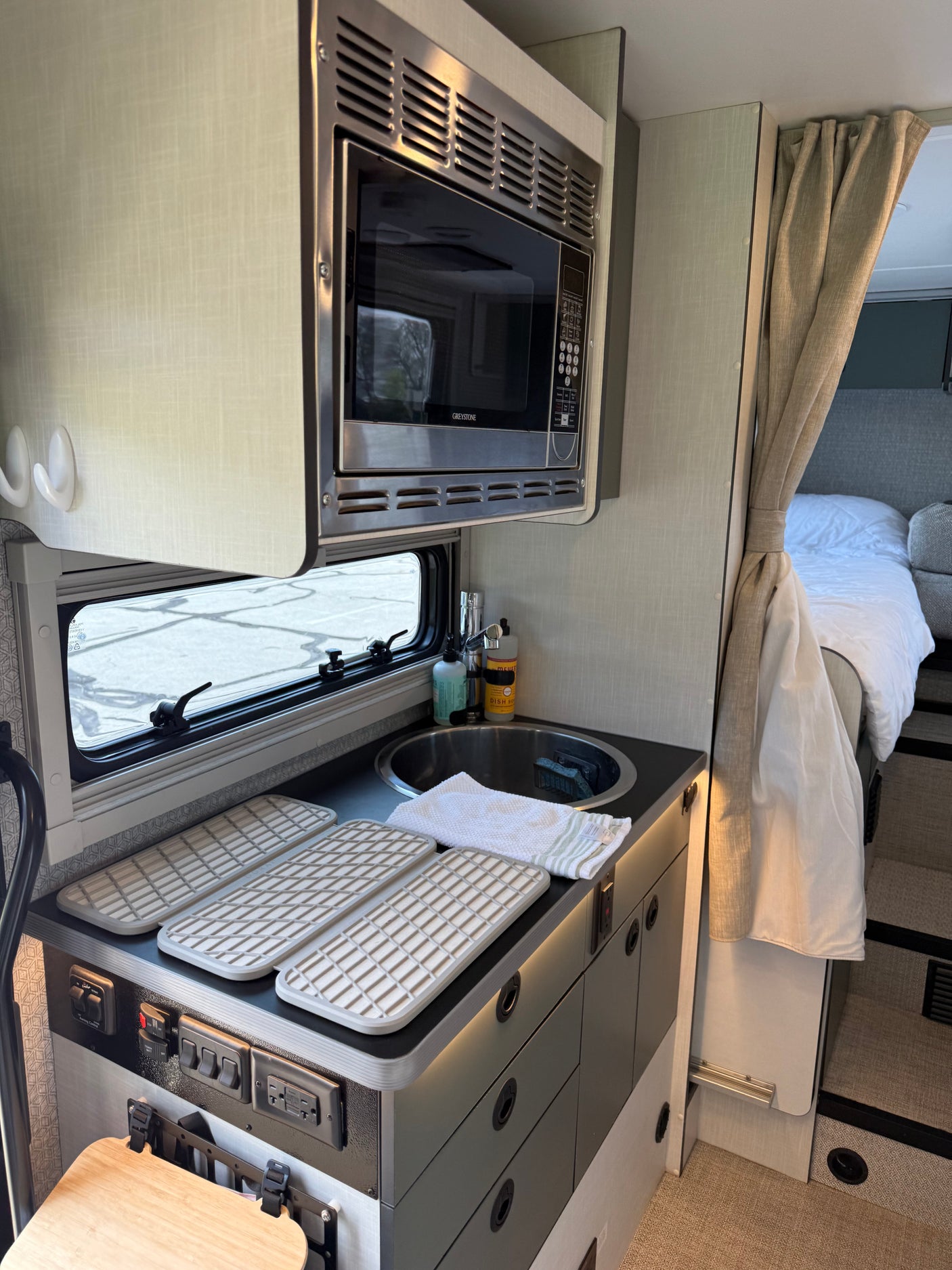 Inside an RV rental: modern kitchen with microwave, sink, drying mats, controls, and a peek into the cozy bed space.