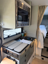Inside an RV rental: modern kitchen with microwave, sink, drying mats, controls, and a peek into the cozy bed space.