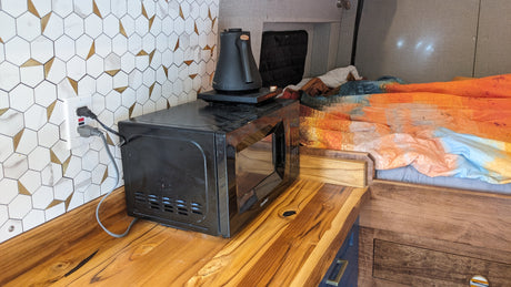 RV rental kitchen setup with microwave, electric kettle, and hexagon tile backsplash next to a cozy bed area.