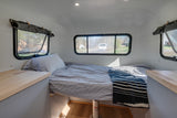 Interior view of a minimalist RV rental: a cozy bed with striped bedding, windows showing an outdoor scene, and light wood counters.