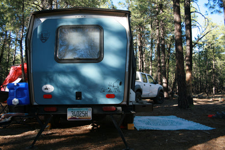 Blue RV rental trailer, tent, and pickup truck nestled in a sunny pine forest campsite. Enjoy outdoor adventures!