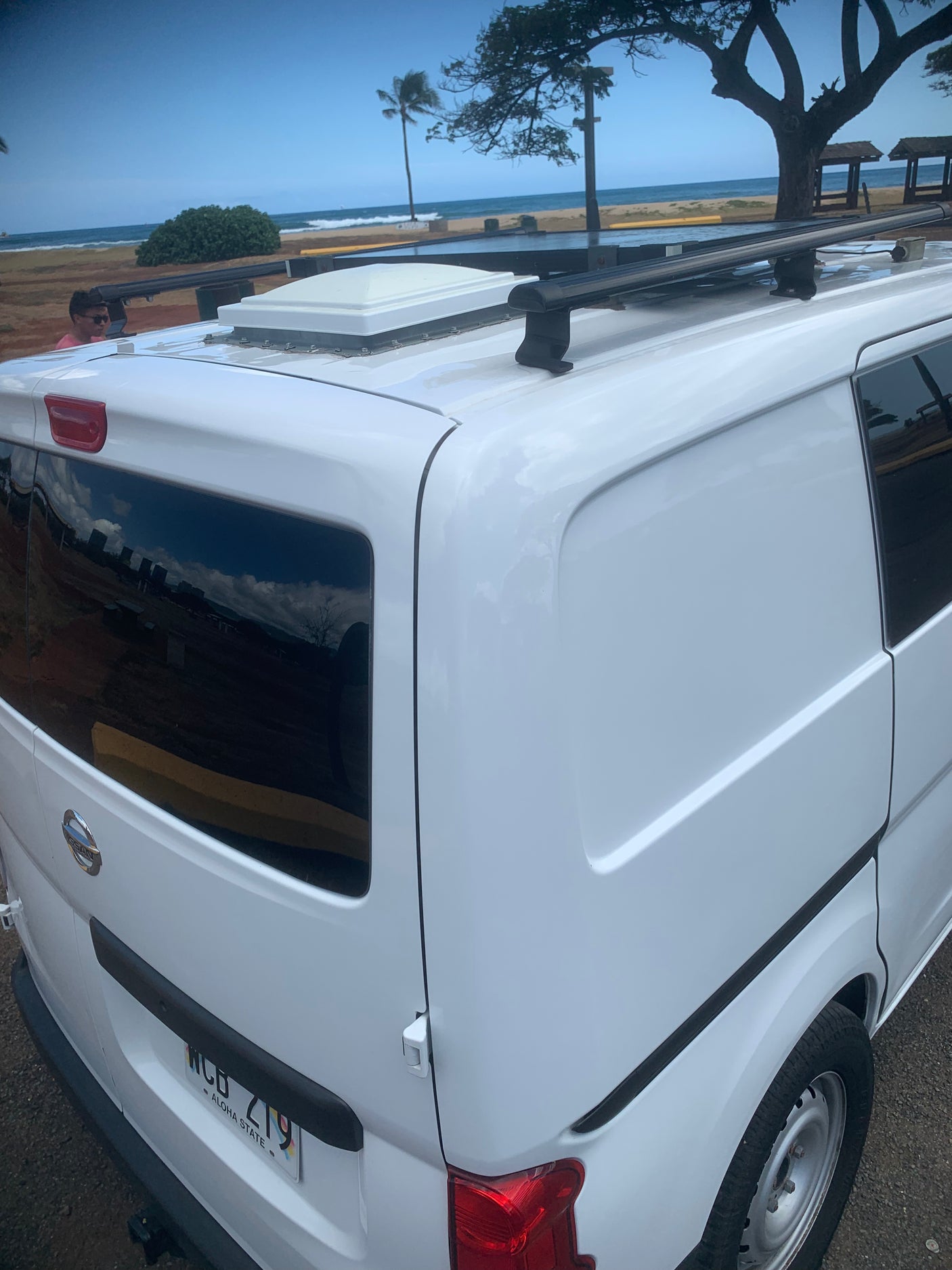White Nissan camper van with roof racks by the beach, reflecting ocean and trees. Perfect RV rental for your coastal adventure.