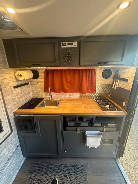 This RV rental's kitchen features a compact design with a sink, stove, wood counters, and clever storage, perfect for mobile cooking.