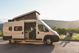 Scenic RV rental getaway: White camper van with pop-top, side door open, couple watching sunset over mountains.
