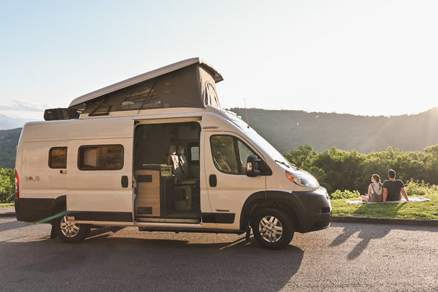 Scenic RV rental getaway: White camper van with pop-top, side door open, couple watching sunset over mountains.