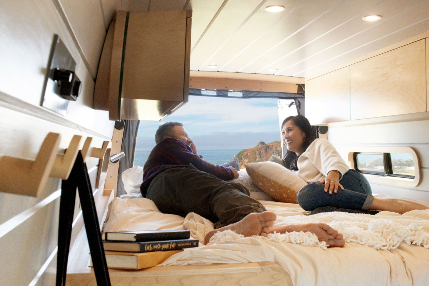 Smiling couple relaxes inside a bright rv rental camper van. Ocean & cliff views seen through the open back. Cozy travel awaits.