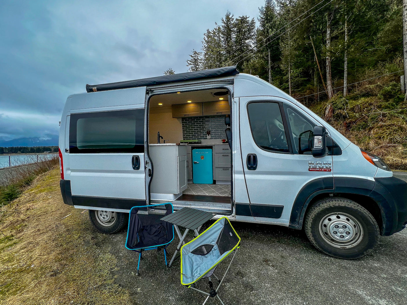 Scenic RV rental van parked by water, open door reveals stylish kitchen & outdoor setup with chairs.