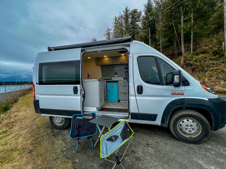 Scenic RV rental van parked by water, open door reveals stylish kitchen & outdoor setup with chairs.