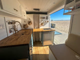 RV rental: Enjoy a bright camper van kitchen with wooden counters and fridge, open to a stunning beach and ocean view.
