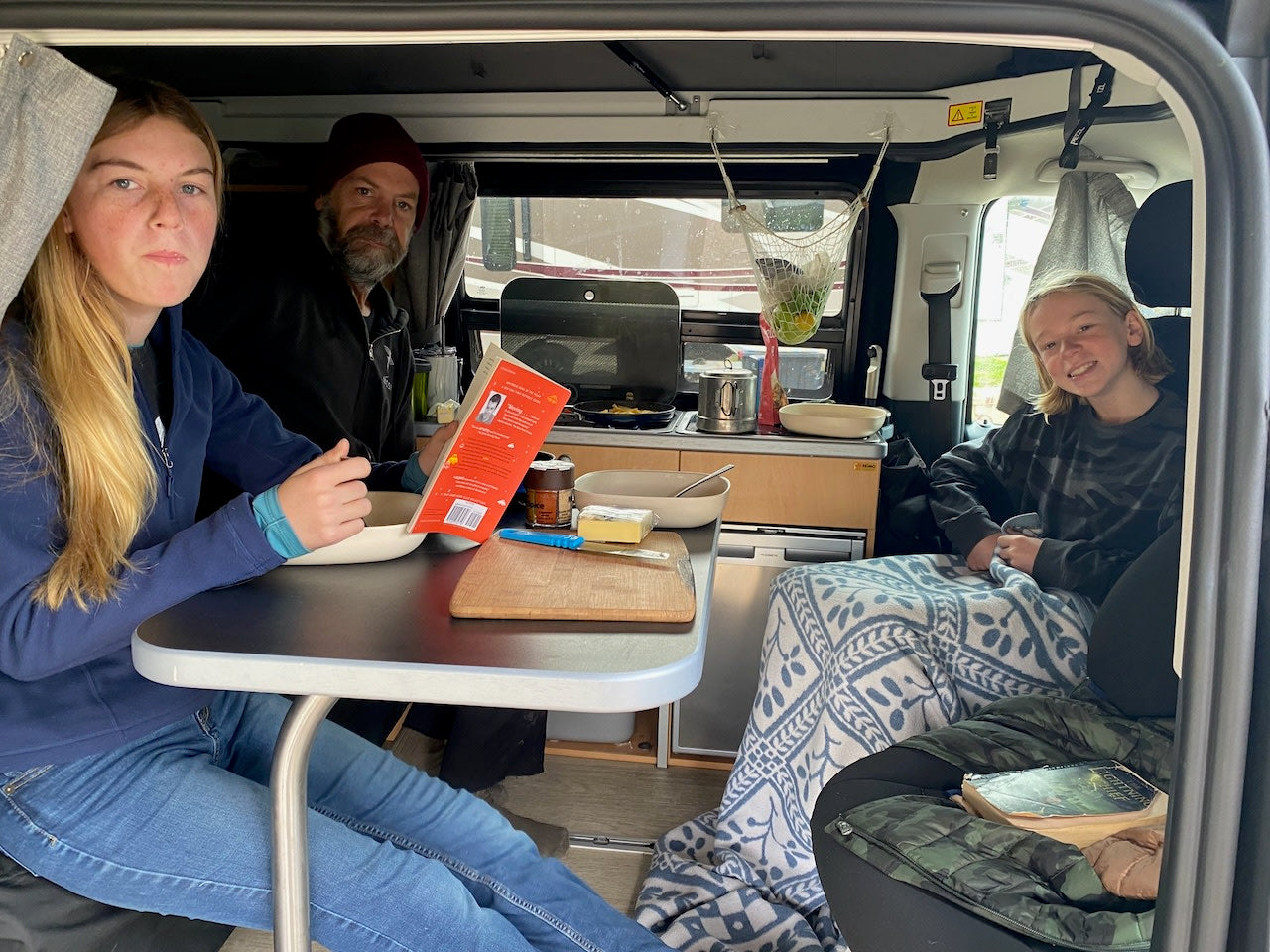 Family inside their RV rental: A young woman looks at the camera, a man reads, and a child smiles from the back. Cozy camper life!