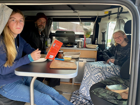 Family inside their RV rental: A young woman looks at the camera, a man reads, and a child smiles from the back. Cozy camper life!