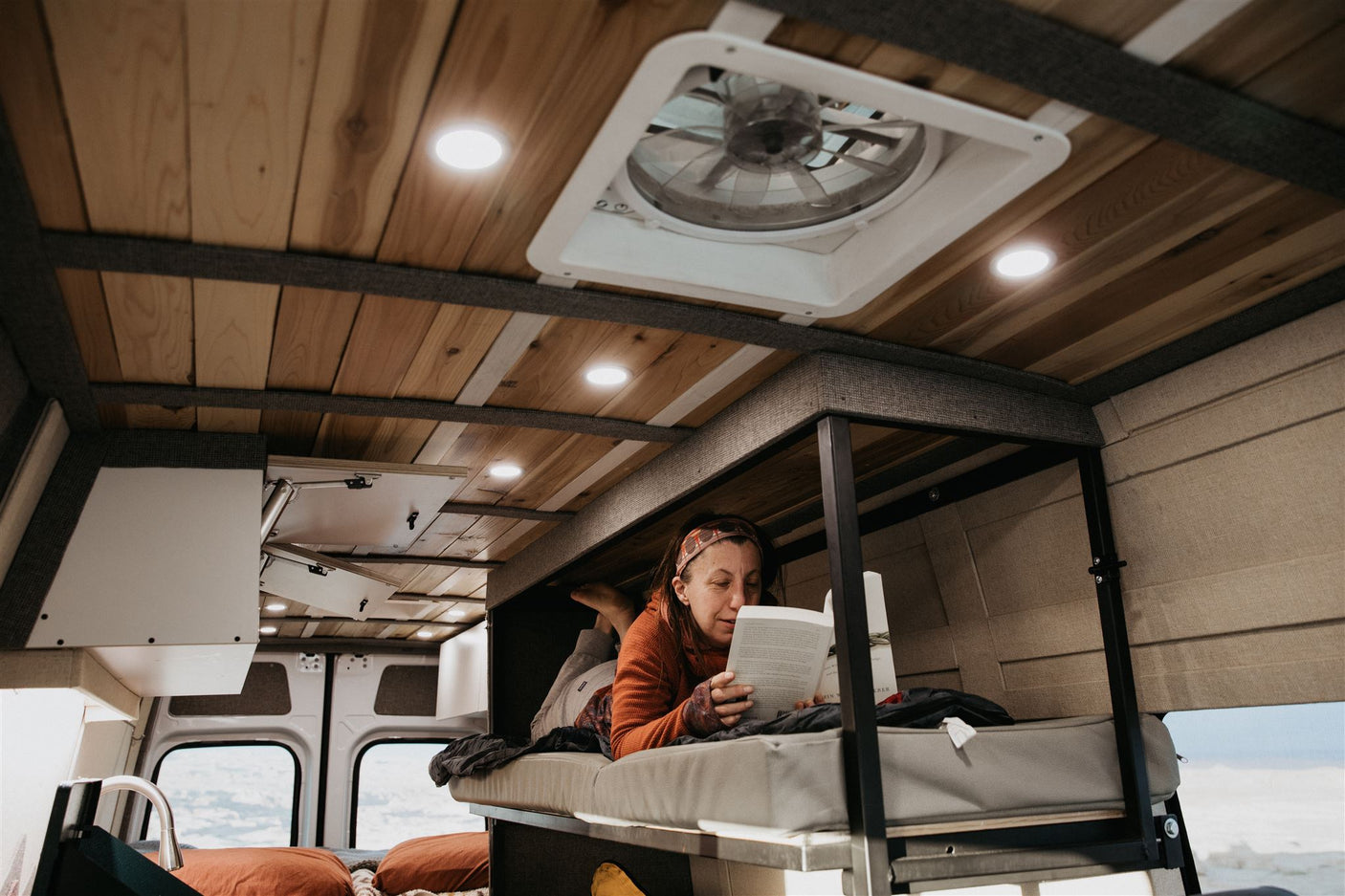 Cozy rv rental interior: woman relaxes reading in a custom camper van bunk, showcasing built-in lights & wooden ceiling. Ideal travel comfort.