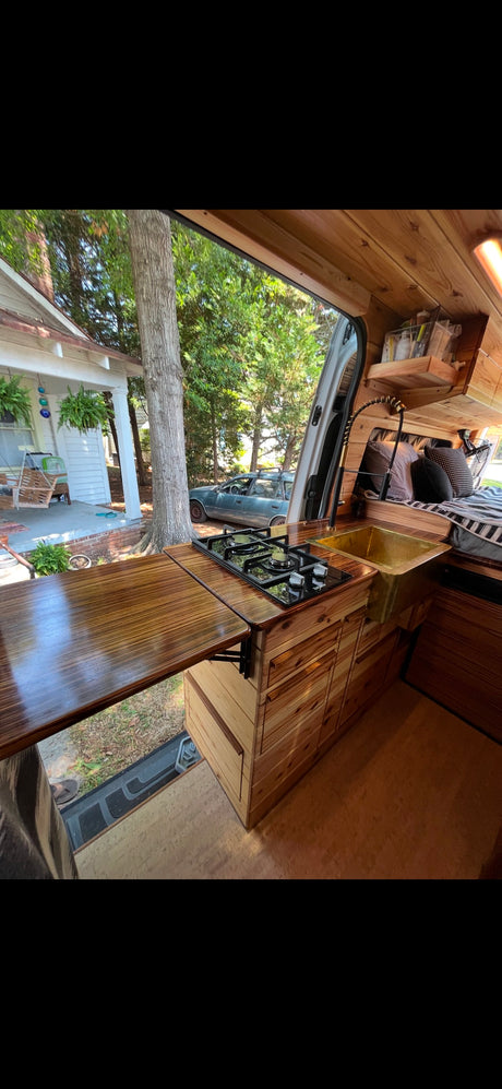 Inside a cozy RV rental: wooden kitchen with stove, golden sink, and a fold-out counter, overlooking a house and yard.