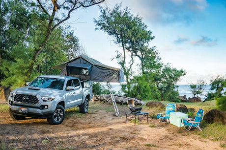 Coastal camping ready! This RV rental features a Tacoma with a rooftop tent, grill, chairs, & cooler on a dirt path near the ocean.