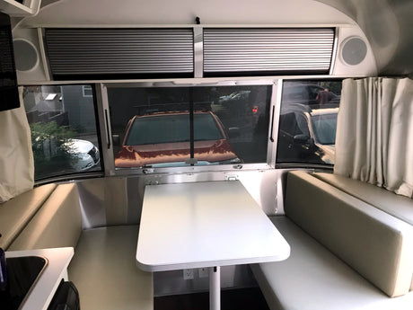 RV rental interior: A modern travel trailer dining area with a white table, light cushions, and large windows revealing parked vehicles outside.
