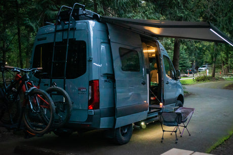 Evening at a wooded campground: a grey Revel RV rental van with bikes attached, awning out, and interior lit for relaxing.
