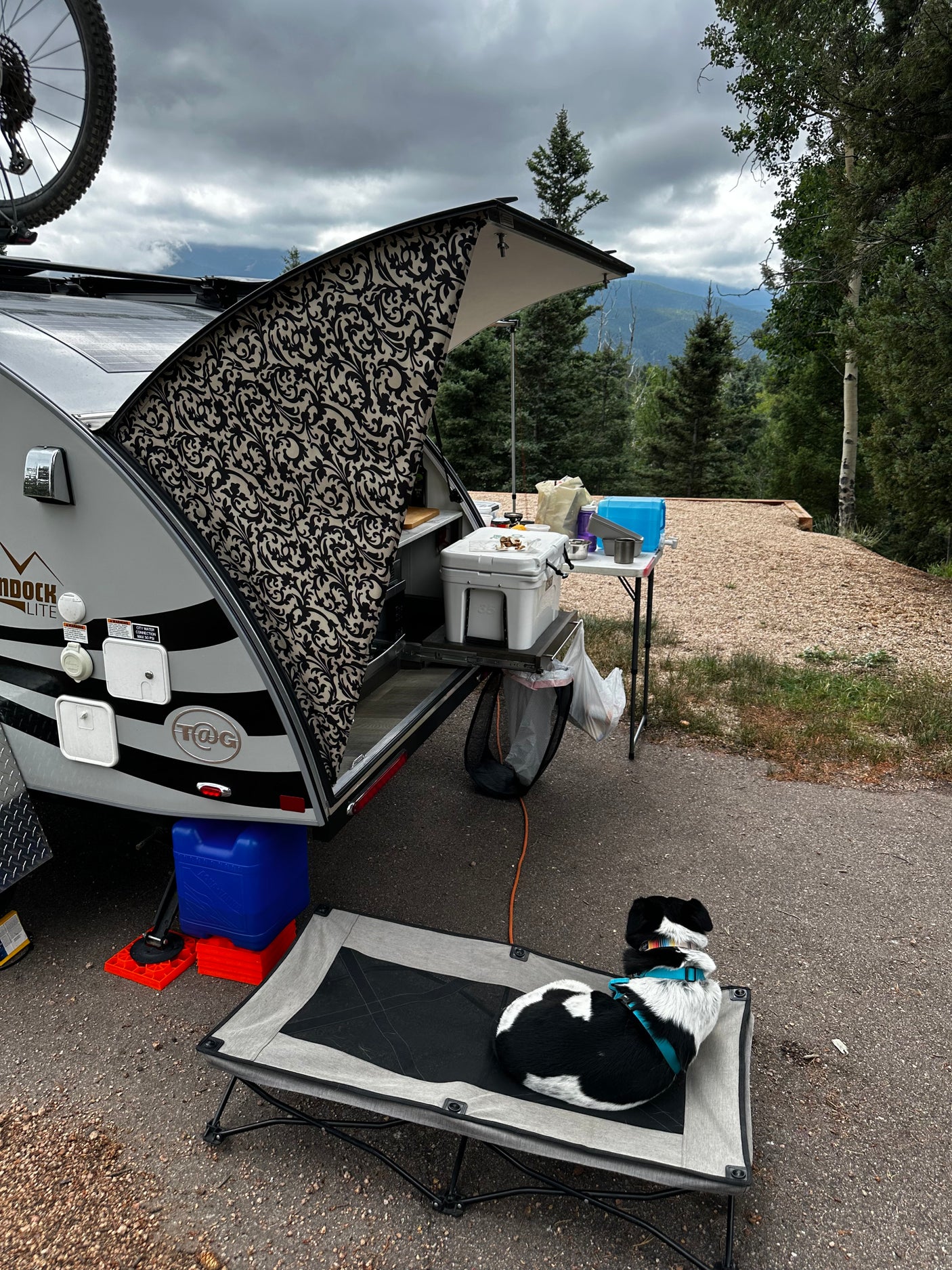 Camping with an RV rental: a compact teardrop trailer's outdoor kitchen and a resting dog. Scenic mountains beyond.