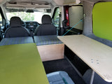 Compact RV rental interior: patterned bed cushions, wooden storage, and a pull-out table. Green mattress on left.
