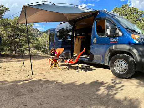 A blue Ram ProMaster RV rental with an awning extends over a girl in a camp chair, enjoying the outdoors.
