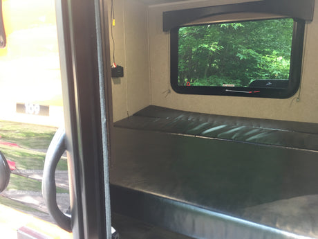 Inside an RV rental, a dark bunk bed faces a window with lush green trees. An exit sign and wall wiring are visible.