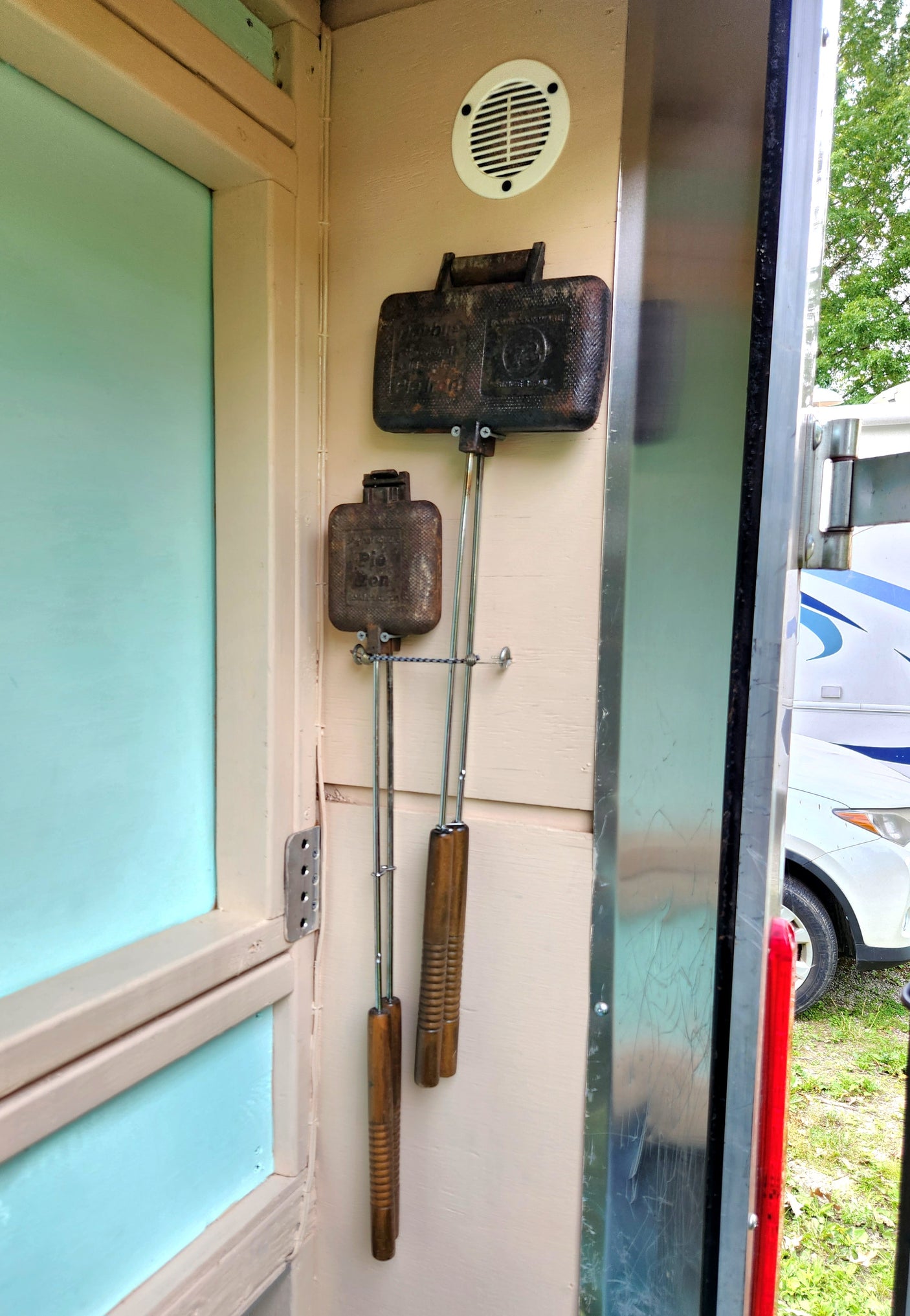 Rustic cast iron pie irons with long wooden handles hang on a light wall inside what looks like an RV rental, set for camping.