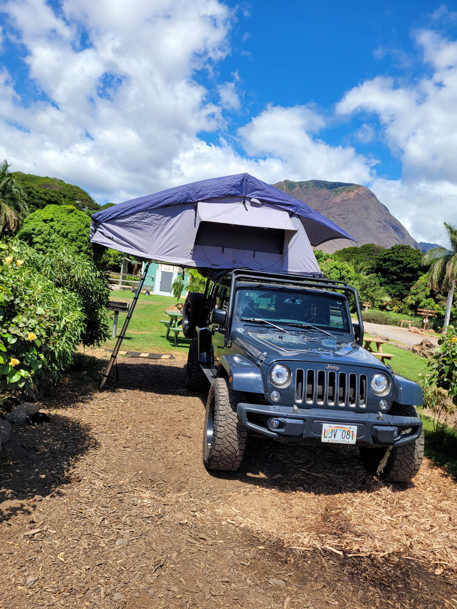 A dark gray Jeep Wrangler with a deployed rooftop tent offers a unique RV rental for camping amidst Hawaii's scenic beauty.
