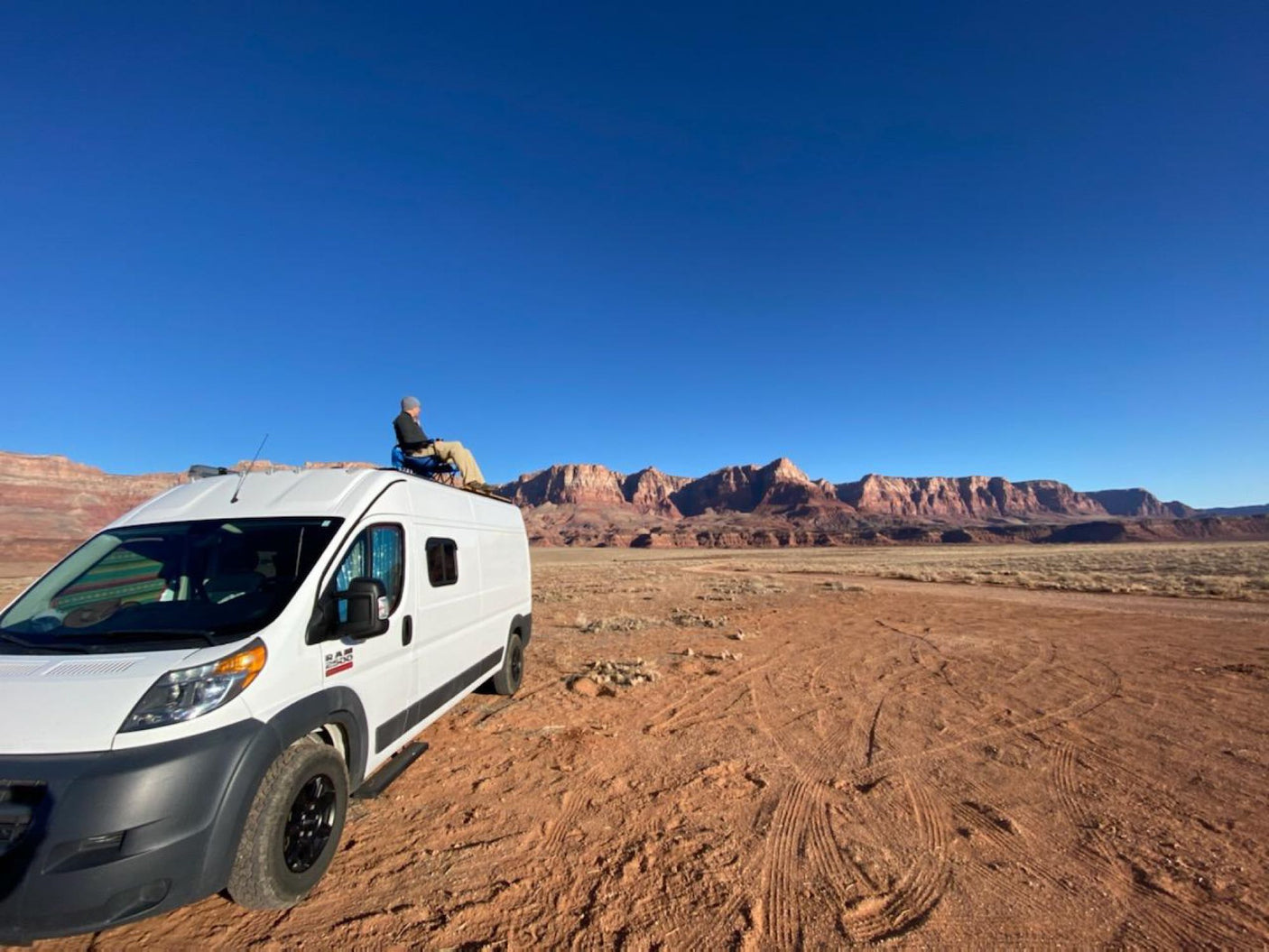A person relaxes on top of an RV rental van, gazing at red desert mountains under a clear blue sky, ideal for remote camping.