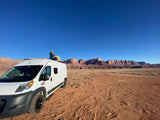 A person relaxes on top of an RV rental van, gazing at red desert mountains under a clear blue sky, ideal for remote camping.