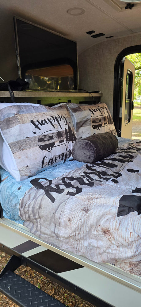 Cozy RV rental bed with 'Happy Camper' wood-grain bedding, a TV, and an open door for refreshing camp views.