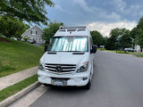 A white Mercedes-Benz Sprinter RV rental van with a sunshade, parked on a residential street under a cloudy sky.