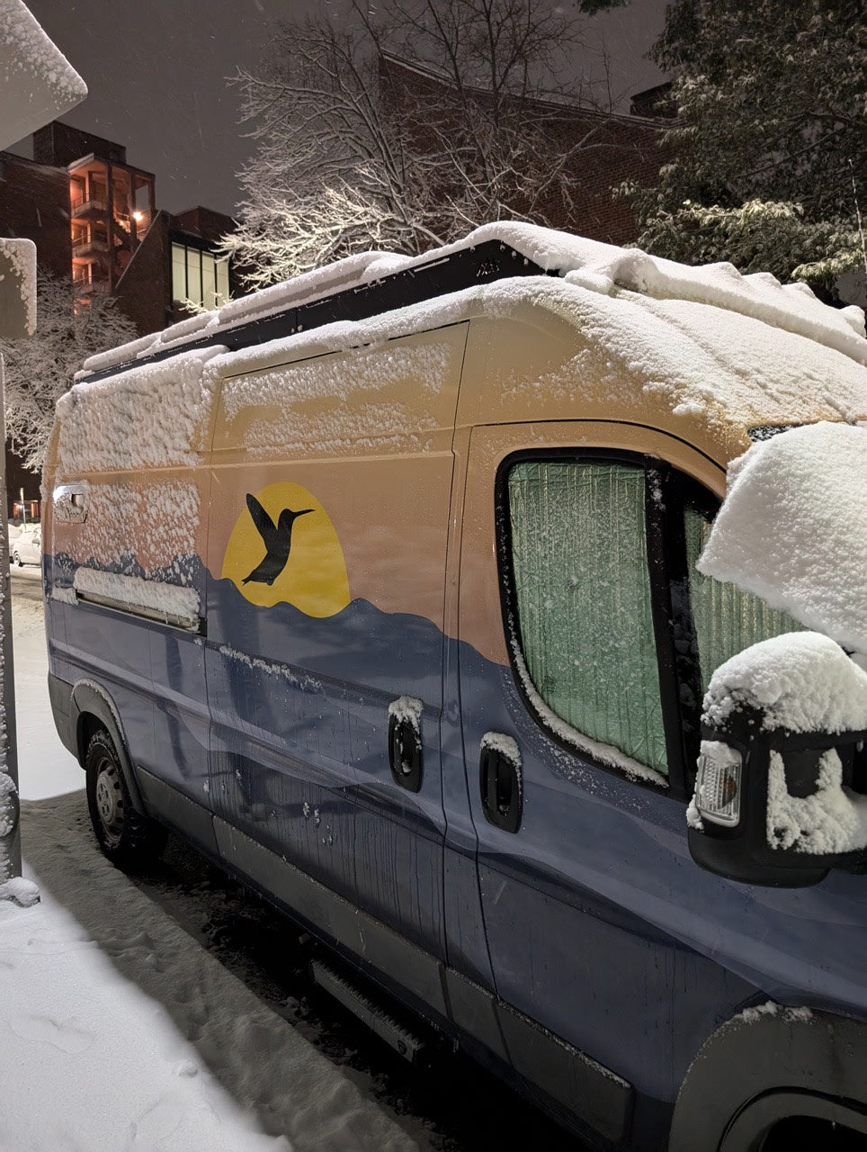 Snow-covered custom camper van (RV rental) at night. Features a hummingbird mural; ready for winter adventures.
