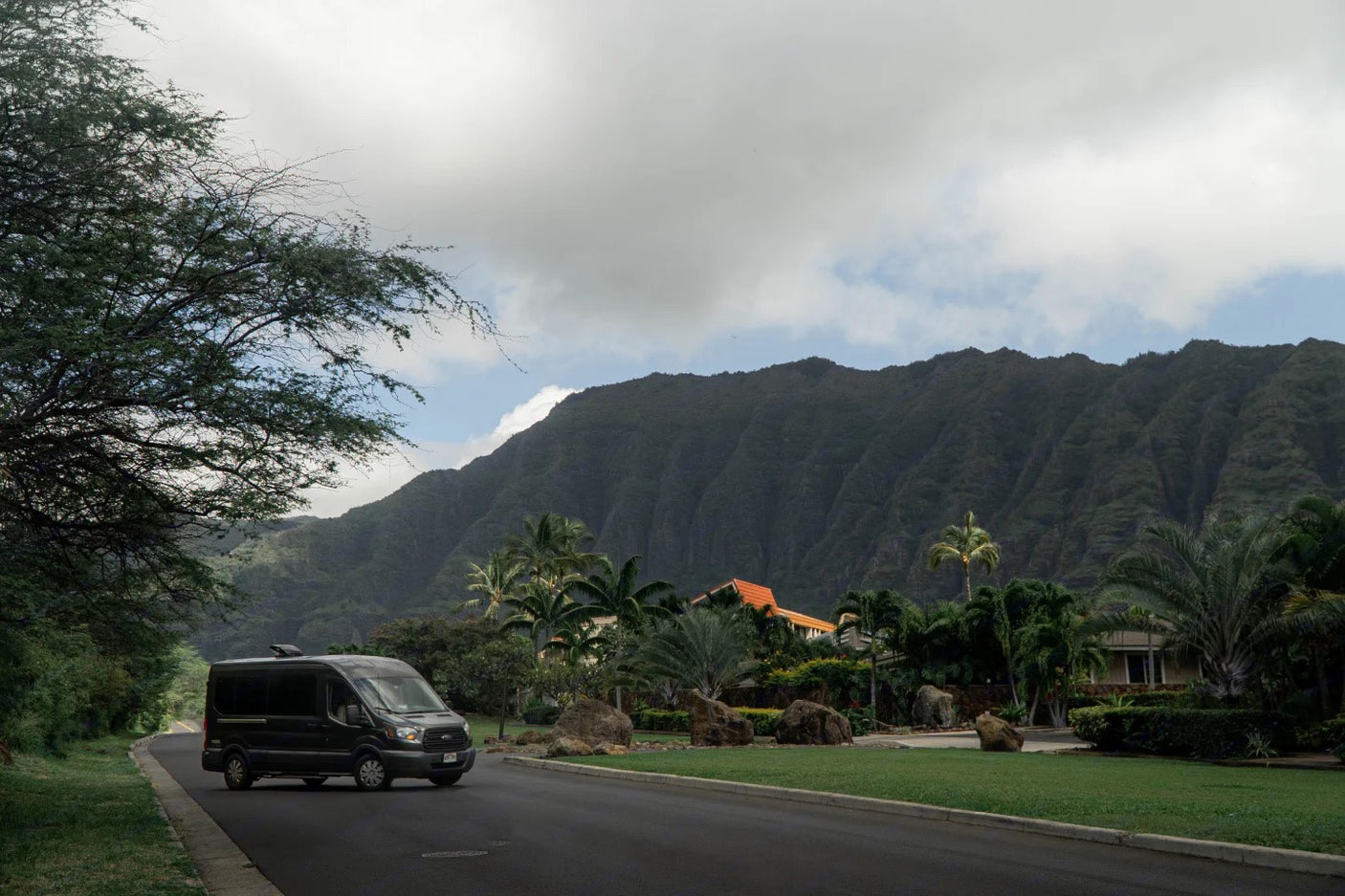 Dark RV rental van on a tropical road, with majestic green mountains and palm trees under a cloudy sky. Scenic travel awaits!