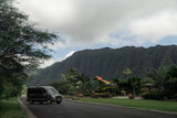 Dark RV rental van on a tropical road, with majestic green mountains and palm trees under a cloudy sky. Scenic travel awaits!