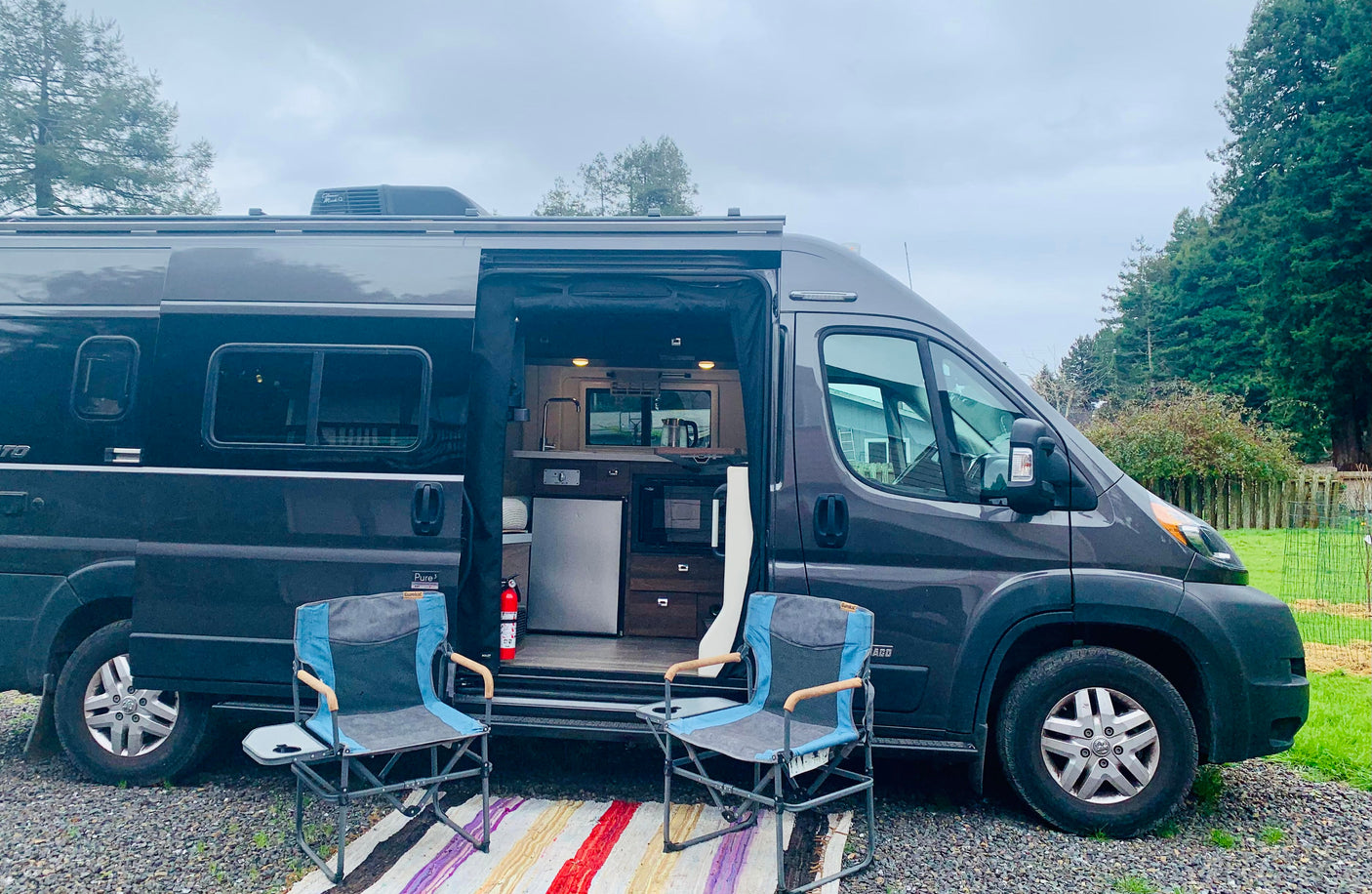 Experience comfort with this dark grey camper van RV rental. Open door shows interior kitchen, two chairs on rug outside.