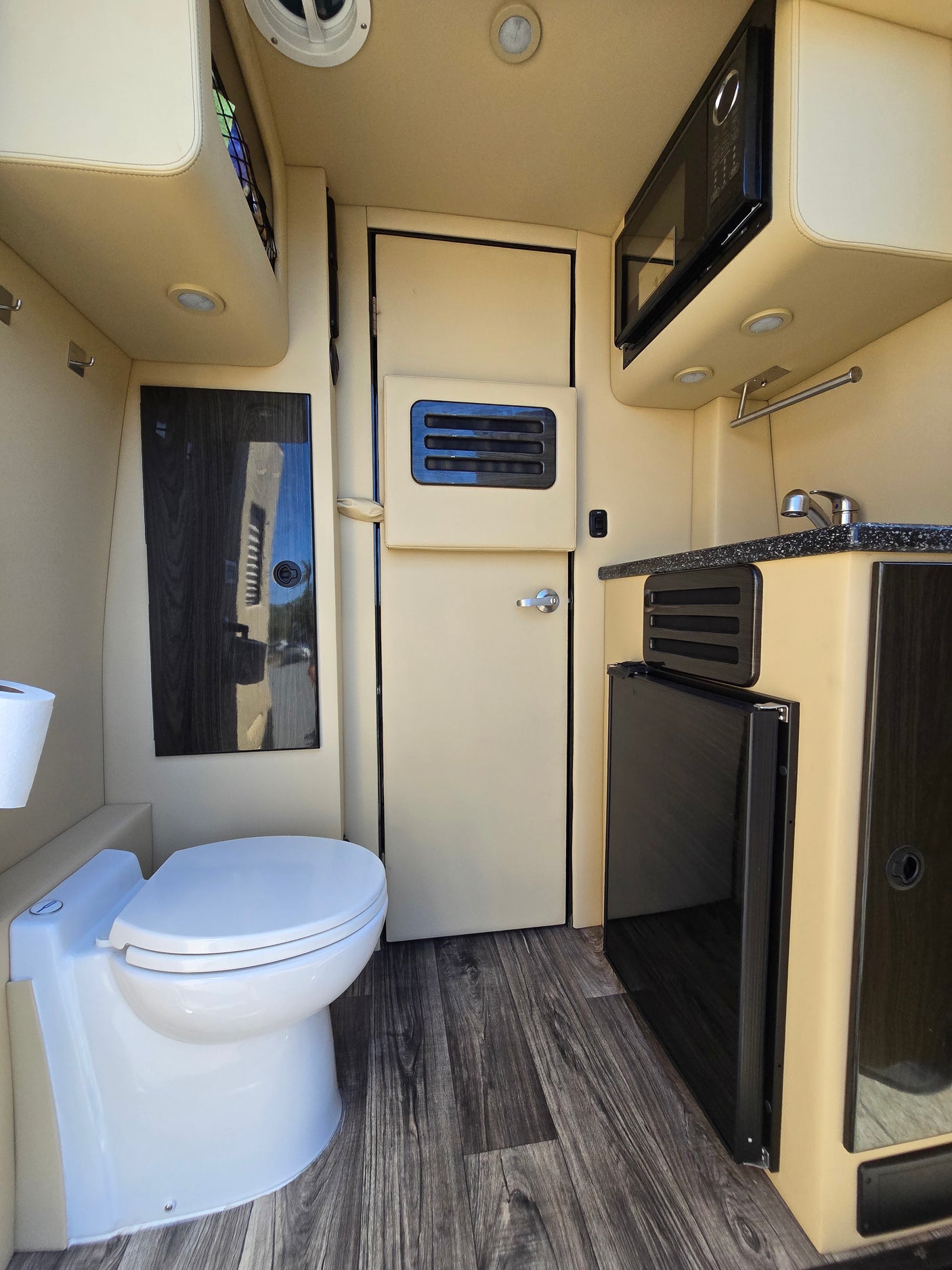 RV rental: Inside a camper van featuring a toilet, kitchenette with sink, microwave, and mini-fridge on wood-look flooring.