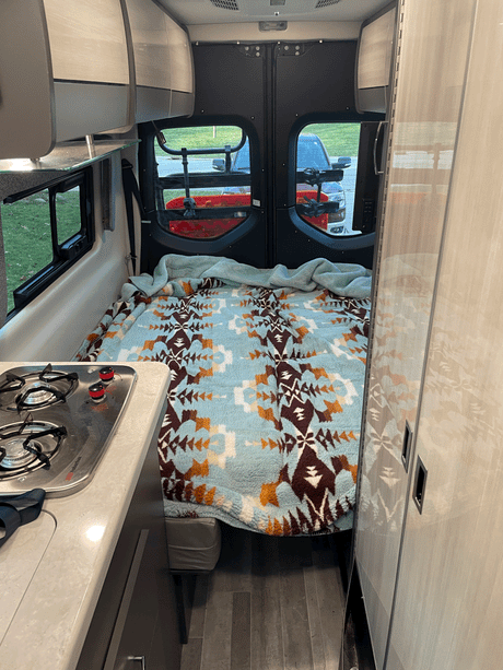 Rv rental camper van interior: cozy bed with patterned blanket, small kitchen with stove, and rear windows.