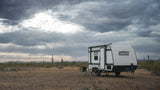 Coleman rv rental in a vast, rocky desert. A travel trailer with chairs set up, under a dramatic, cloudy sky with sun rays.