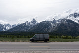 Stunning landscape with a grey RV rental van on a road, backed by impressive snow-capped Grand Teton mountains and forest.
