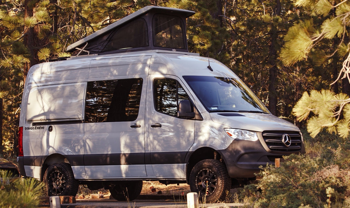 Light grey Mercedes Sprinter camper van with pop-top roof, parked among trees. Ideal RV rental for your next outdoor adventure.