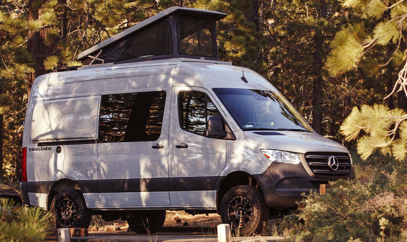 Light grey Mercedes Sprinter camper van with pop-top roof, parked among trees. Ideal RV rental for your next outdoor adventure.