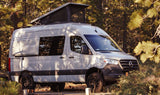 Light grey Mercedes Sprinter camper van with pop-top roof, parked among trees. Ideal RV rental for your next outdoor adventure.