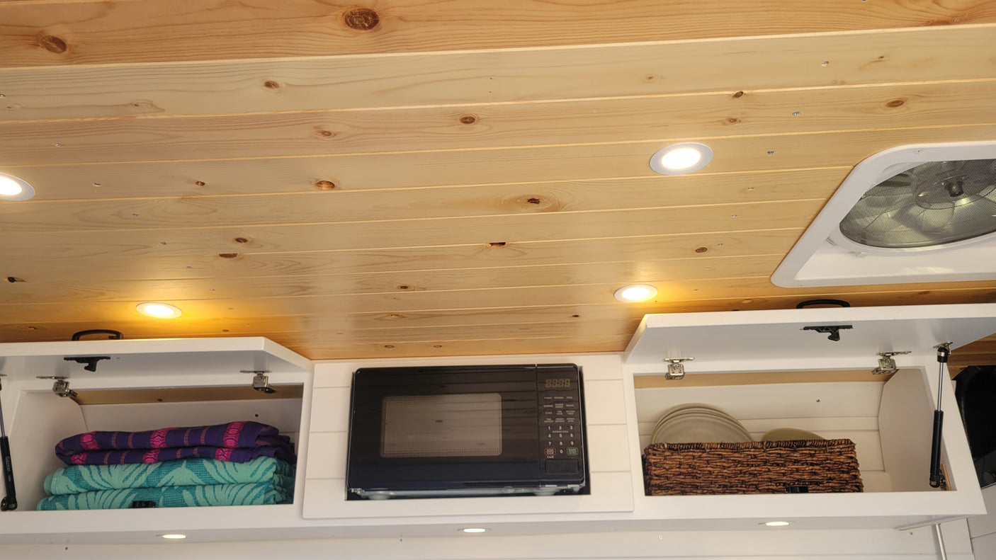 RV rental's overhead storage: wood ceiling, bright lights, microwave, and open cabinets with towels and dishes.