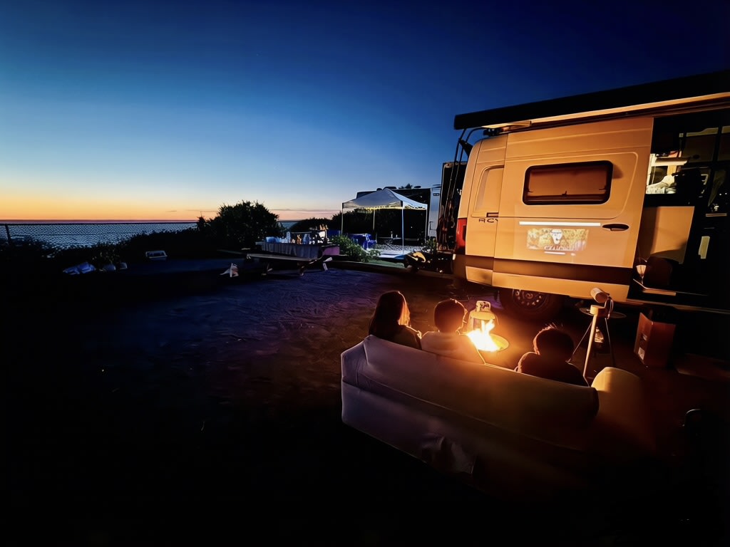 RV rental family fun: An outdoor movie projected on the van at sunset, by the ocean. Kids and adults cozy by the fire.