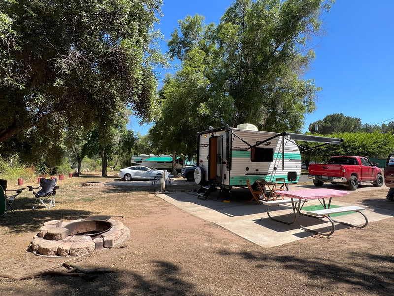 Sunny campsite featuring a retro-style RV rental, picnic table with red checkered cloth, and stone fire pit under shaded trees. — photo 13