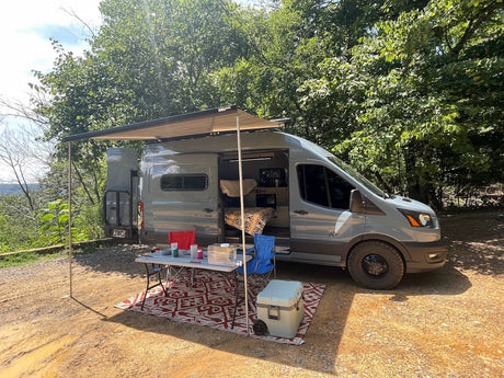 This gray Ford Transit camper van RV rental features an extended awning, table, & rug setup for a scenic outdoor adventure.