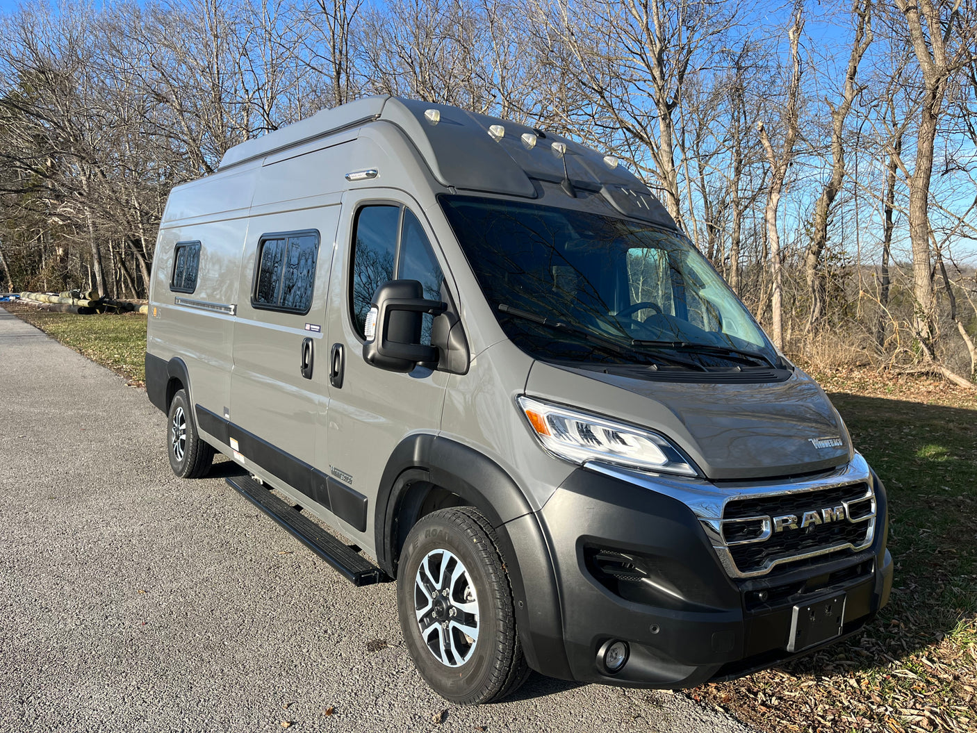 A modern gray Winnebago Solis camper van, a prime RV rental choice, parked outdoors on a bright, clear day among bare trees.