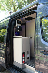 Modern RV rental interior: kitchen with sink, control panel, fire extinguisher, and outlets visible through the open camper van door.