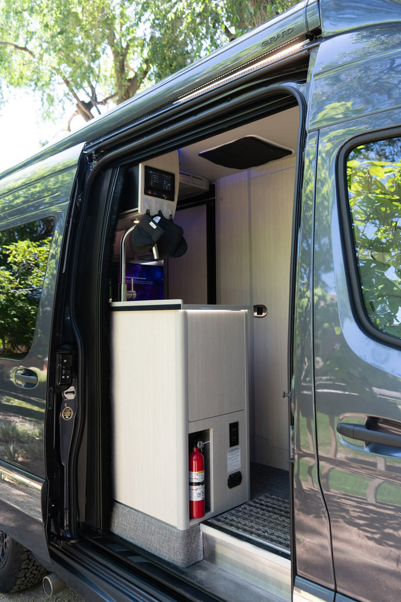 Modern RV rental interior: kitchen with sink, control panel, fire extinguisher, and outlets visible through the open camper van door. — photo 9